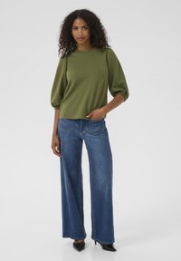 Oliven grøn tekstureret top med pufærmer, parret med bredbenede blå denimjeans og sorte spidse hæle. Enkel og struktureret design.