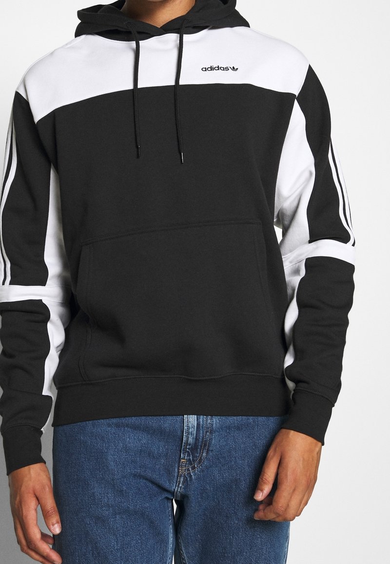 Svart och vit Adidas-huvtröja med dragsko i huvan, färgblockdesign och tre vita ränder på ärmarna. Har en framficka.