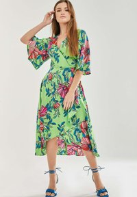 Green wrap dress with vibrant floral print in pink and blue. Features bell sleeves, an asymmetric hem, and a smooth fabric texture.
