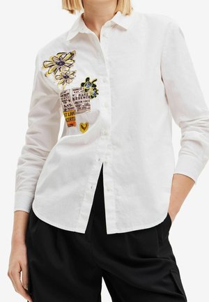 Witte overhemd met knopen en kleurrijke bloemen- en tekstapplicaties op de linkerborst, gecombineerd met een zwarte broek, model met hand in de zak.