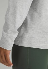 Jasnoszary, długi rękaw, top o teksturowanym wykończeniu, z przeszytym dołem i małym logo z boku, w zestawie z ciemnozielonymi legginsami.