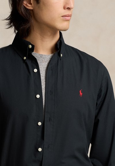 Moški v črni srajci z gumbi čez sivo majico z okroglim izrezom, na prsih pa rdeč vezen konjiček.