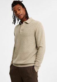 Ljusbeige långärmad polo skjorta med texturerad stickning, tre-knapps knäppning och ribbade ärmslut och nederkant, i kombination med mörkbruna byxor.