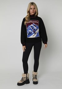 Donna che indossa un maglione nero con una scena di sci in montagna, leggings neri e stivali in shearling beige, in piedi contro uno sfondo semplice.
