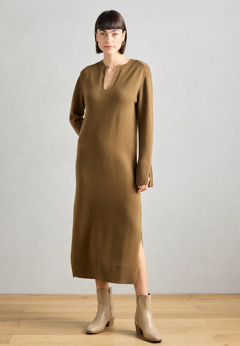 Lungo vestito marrone a maglia con scollatura a V, dettagli a coste e aperture laterali, abbinato a stivali beige fino alla caviglia su uno sfondo di pavimento in legno.