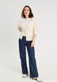 Helle beige gestrickte Strickjacke mit V-Ausschnitt und Knopfverschluss, über einem weißen Spitzenoberteil getragen. Kombiniert mit weiten blauen Jeans und Turnschuhen.