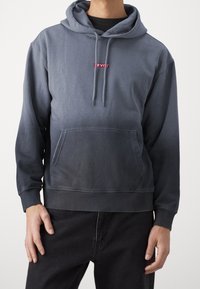 Felpa con cappuccio grigia con sfumatura che diventa più scura verso il fondo, dotata di una toppa rossa con logo sul petto e di una tasca frontale.