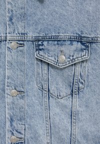 Section d'une veste en denim bleu clair montrant des boutons en métal et une poche à rabat avec fermeture à bouton sur un tissu texturé.