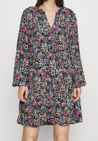 Kukallinen mekko, jossa on musta tausta ja vaaleanpunaisia, sinisiä ja vihreitä kukkia. Pitkät hihat ja V-päinen muotoilu tuovat lisää yksityiskohtia.