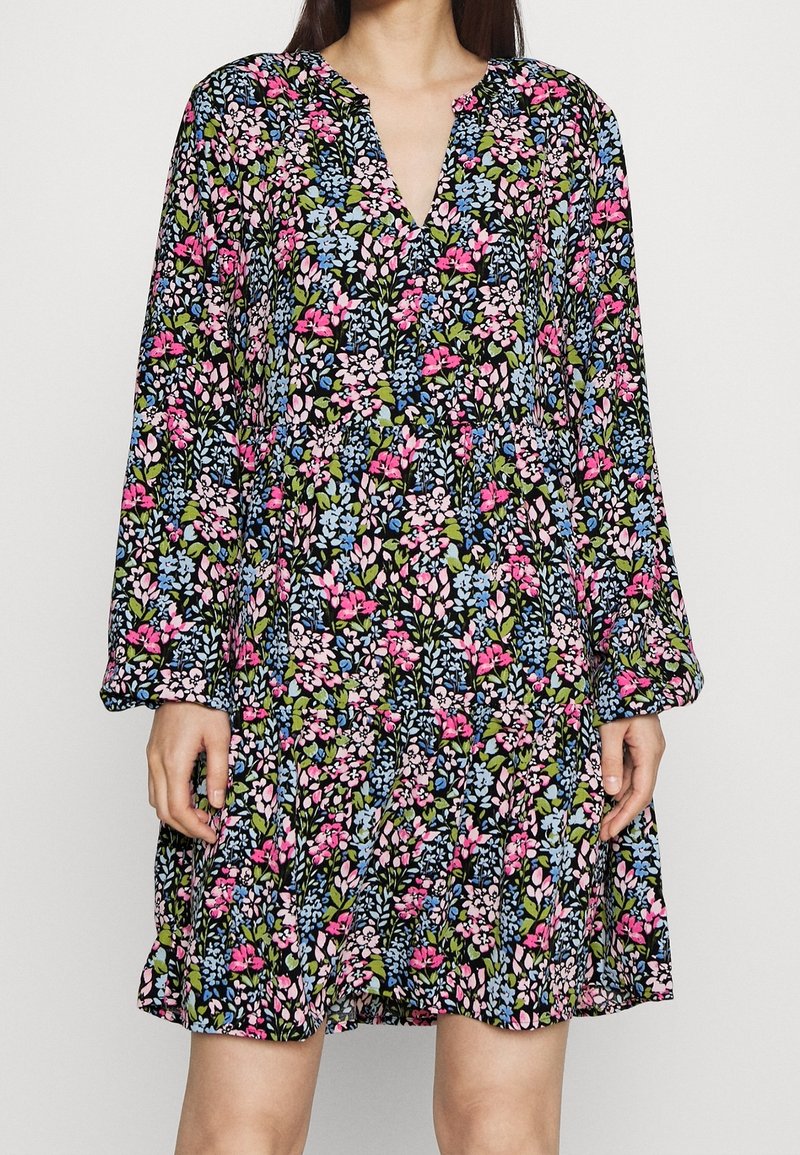 Kukallinen mekko, jossa on musta tausta ja vaaleanpunaisia, sinisiä ja vihreitä kukkia. Pitkät hihat ja V-päinen muotoilu tuovat lisää yksityiskohtia.