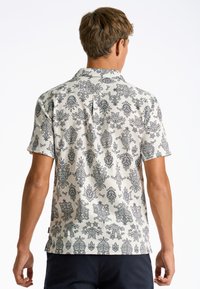 Camisa de manga corta en crema con intrincados patrones florales negros. Cuenta con un cuello con solapa y un pliegue central en la parte posterior para facilitar el movimiento.