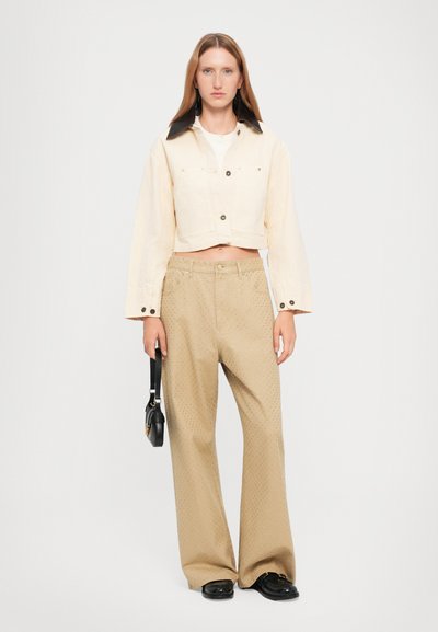 Jachetă din denim crem, scurtă, cu guler negru, asortată cu pantaloni largi bej, texturați, cu mici accente de model. Ținând o geantă mică neagră.