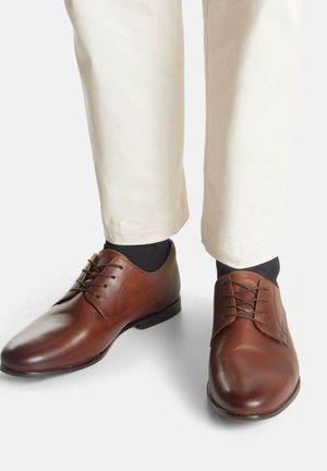 Zapatos de vestir de cuero marrón con un acabado liso, punta redonda y diseño de cordones con cinco ojales. Combinados con pantalones beige y calcetines oscuros.