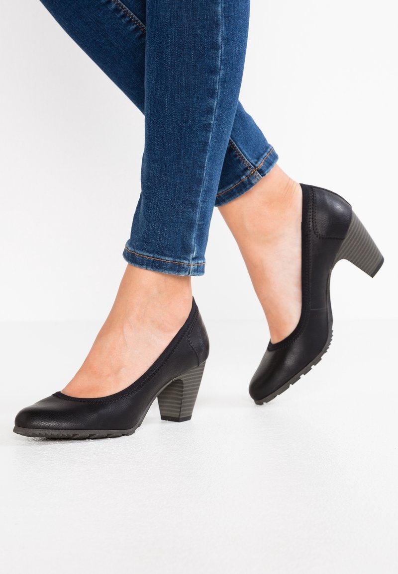 Personne portant un jean skinny bleu et des escarpins en cuir noir à talons moyens avec semelles texturées, debout sur une surface blanche.