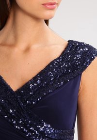 Marindress med en paljettprydd axeldesign, mjuk tyg, och en V-ringning som framhäver en texturerad, skimrande övre detalj.