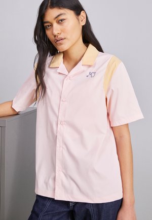 Młoda kobieta opiera się o szary mebel, ubrana w różową koszulę z krótkim rękawem z beżowym kołnierzykiem i niebieskimi haftowanymi inicjałami.