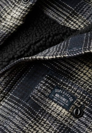 Camisa de franela a cuadros en blanco y negro con un interior suave y peludo, con un botón negro y una etiqueta que dice "Superdry Co."