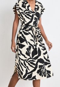 Femme portant une robe beige mi-longue avec de grands imprimés floraux abstraits noirs et une ceinture nouée à la taille, se tenant devant un fond uni.