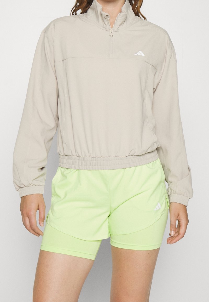 Personne portant un haut beige à manches longues zippé et un short de sport superposé vert clair, debout devant un fond uni.