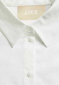 Camicia bianca con colletto classico, realizzata in tessuto morbido. Presenta un'etichetta con la scritta "JJXX" sotto il colletto e bottoni semplici.
