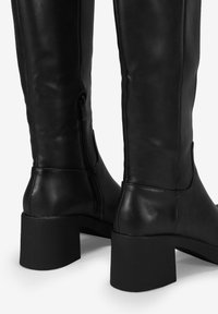 Botas de cuero negro por encima de la rodilla con un acabado liso, tacón cuadrado y detalle de cremallera lateral. Diseñadas para una silueta ajustada.