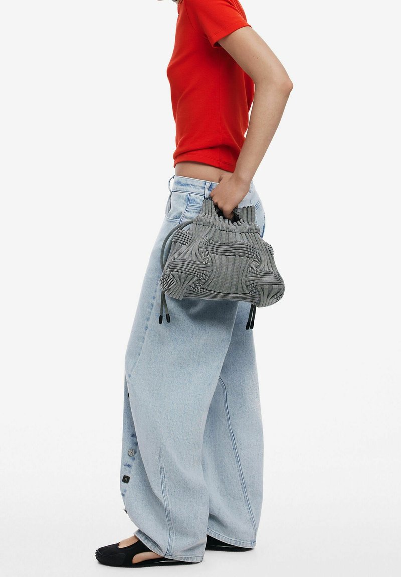 Personne portant une chemise rouge à manches courtes et un jean large bleu clair, tenant un sac à main gris texturé, debout devant un fond blanc uni.