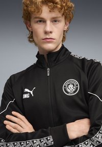 Czarna kurtka sportowa z zamkiem błyskawicznym, haftowanym logo Manchester City, białym logo Pumy oraz wzorzystymi akcentami materiałowymi na rękawach.