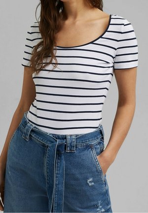 Kobieta ubrana w biały top z krótkim rękawem z granatowymi poziomymi paskami oraz niebieskie dżinsy z wysokim stanem, z materiałowym paskiem i ręką w kieszeni.