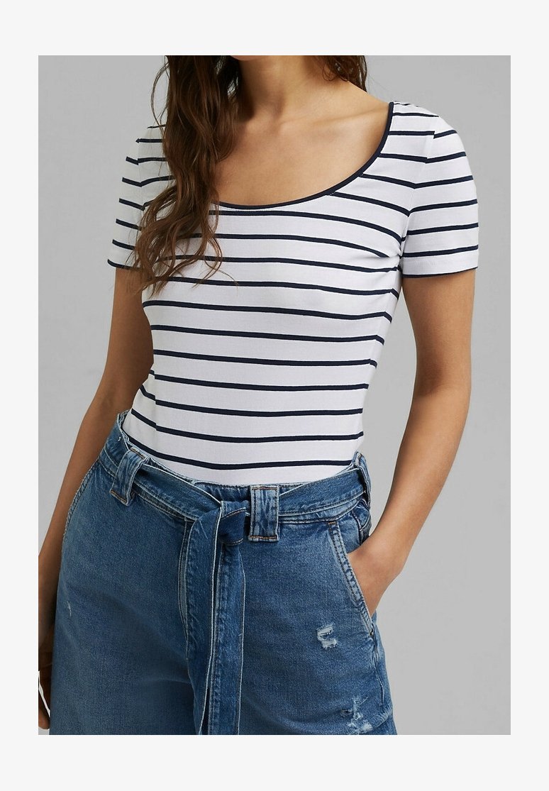 Vrouw draagt een wit T-shirt met korte mouwen en marineblauwe horizontale strepen, gecombineerd met een hoge taille blauwe spijkerbroek met een stoffen riem en haar hand in de zak.