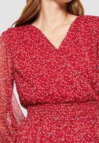 Mujer vistiendo un vestido floral rojo con mangas largas y transparentes y una cintura entallada, mostrado de cuello a cintura.