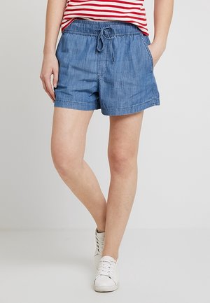Femme portant un short bleu en jean avec cordon, un haut rayé rouge et blanc, et des baskets blanches, debout avec une jambe croisée sur l'autre.