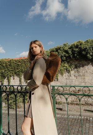 Mochila de ante marrón con parte superior redondeada, llevada al hombro; modelo con un trench beige, contra un fondo de exuberante vegetación.