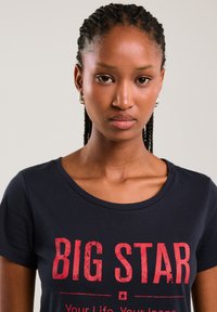 Moteris su pintais plaukais dėvi juodą marškinėlį su ryškiu raudonu užrašu „BIG STAR“, žvelgianti į priekį priešais paprastą šviesią foną.