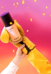 Una mano in camicia bianca tiene un guanto da boxe giallo che afferra una bottiglia di fondotinta KIKO Milano Unlimited su uno sfondo rosa-arancio.