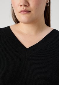 Svart ribbad stickad tröja med V-ringning. Texturen är mjuk och tydlig, vilket framhäver det vävda mönstret och de rena linjerna.