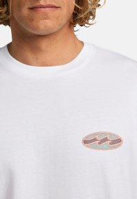Hvid bomulds t-shirt med et rundt, flerfarvet logo med bølgede linjer i brun og teal, placeret på venstre side af brystet.