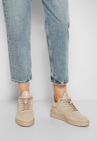 Beżowe zamszowe sneakersy o teksturowanym wykończeniu, z płaskimi sznurowadłami, okrągłym noskiem i niskoprofilową gumową podeszwą. W komplecie z jasnoniebieskimi dżinsami.