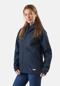 Młoda kobieta z długimi włosami, ubrana w granatową kurtkę na zamek i niebieskie jeansy, stojąca na tle gładkiego jasnego tła.