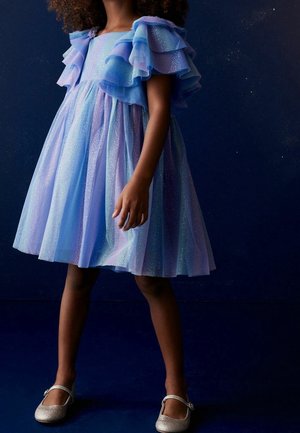 Vestido azul claro y lavanda con mangas con volantes, hecho de tela transparente con toques de purpurina, de capas y hasta la rodilla. Zapatos planos plateados.