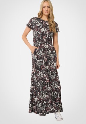 Blonde vrouw die een zwarte maxi-jurk met korte mouwen draagt met een wit en rood paisleypatroon, handen in de zakken, witte sneakers.