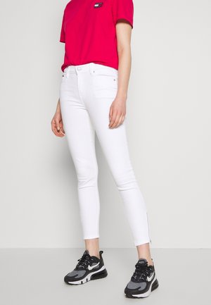 Pantalones vaqueros blancos ajustados con una textura suave, que cuentan con un botón frontal, cierre de cremallera y ligeros cremalleras en los tobillos. Combinados con una camiseta rosa y zapatillas negras.