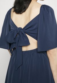 Granatowa sukienka z głębokim dekoltem w serek na plecach, wiązaniem w talii i falbaniastymi rękawami. Miękka tkanina z zwiewnym krojem i marszczonym detalem w pasie.