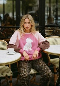 Růžový svetr s hnědými rukávy obsahuje bílou zvířecí grafiku a text "V ZÁBAVĚ". Doplněno leopardími kalhotami na stolku v kavárně.