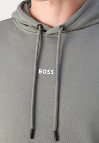 Felpa grigia con cappuccio e tasca a marsupio, caratterizzata da spesse coulisse e un logo bianco "BOSS" sul petto. Tessuto morbido.