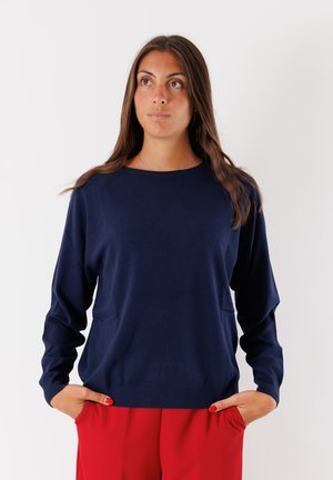 Maglione blu navy con collo a giro, maniche lunghe e orlo a coste. Indossato sopra dei pantaloni rossi, realizzati in un tessuto morbido e texture.