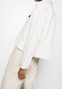 Vit cordjacka med en kortad design, med knappstängning och sidofickor; i kombination med ljusbeiga byxor.