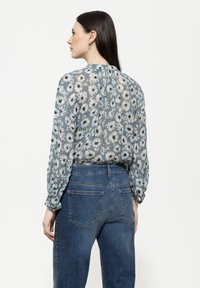 Luftige, durchsichtige Bluse mit blauem Hintergrund und weißem Gänseblümchenmuster, langen Ärmeln und einem Knopfdetail am Rücken, kombiniert mit dunkelblauen Jeans.