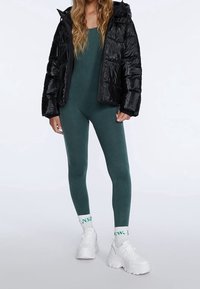 Svart dunjacka med glansig textur över en mörkgrön baddräkt. Vita robusta sneakers och mönstrade ankelsockor fulländar looken.