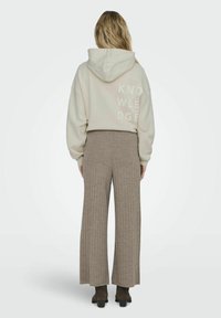 Lichtbeige hoodie met witte bloklettertekst "KENNIS" op de achterkant, gecombineerd met een gestructureerde bruine broek met wijde pijpen.
