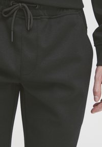 Gros plan sur un pantalon de jogging noir à cordon de serrage porté à la taille et à la cuisse supérieure d'une personne, avec une main visible à côté du pantalon.